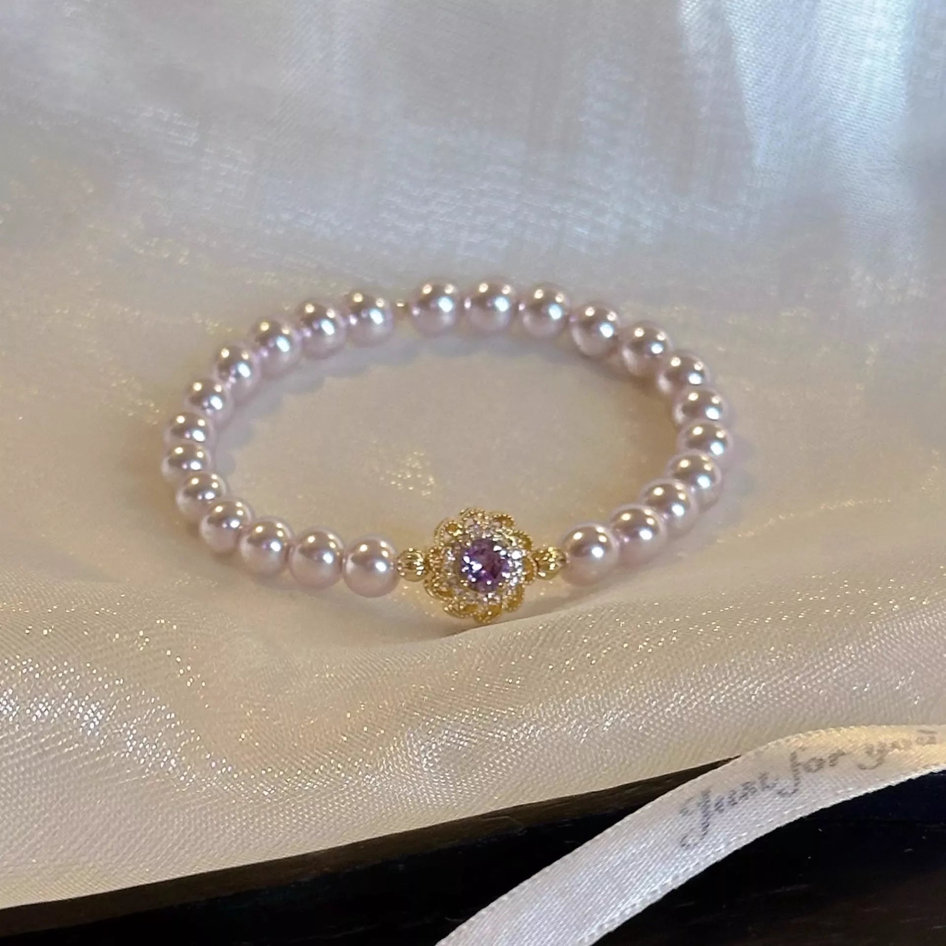 Gold-plated pearl bracelet laid flat: round gold floral charm with purple center on pearl band