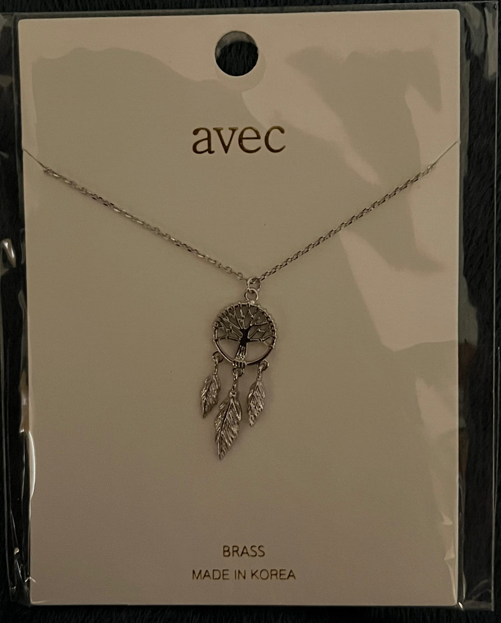 A silver-toned dreamcatcher necklace by the brand avec, displayed on its original cream-colored backing card. The necklace features a circular tree-of-life pendant with dangling feathers.