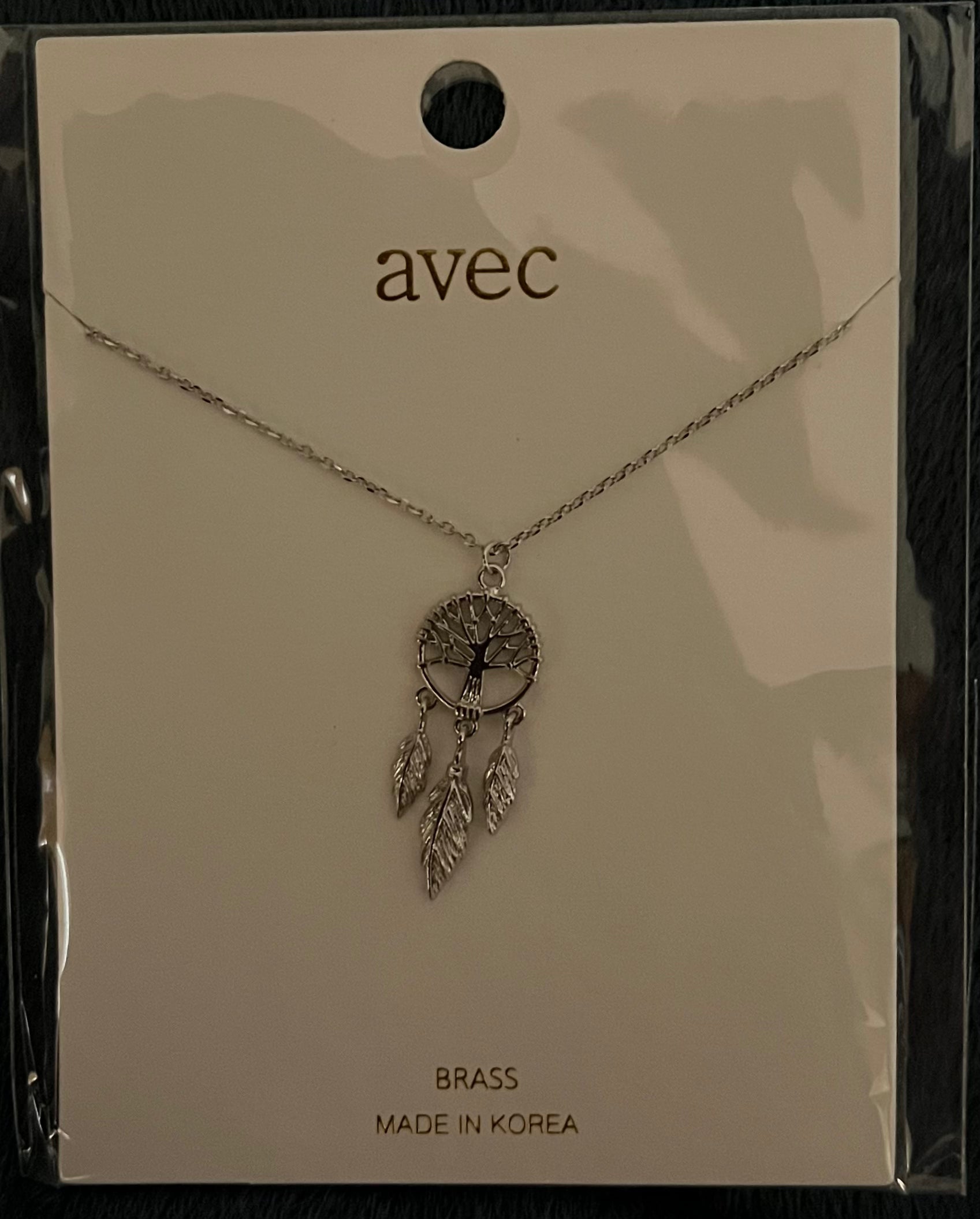 A silver-toned dreamcatcher necklace by the brand avec, displayed on its original cream-colored backing card. The necklace features a circular tree-of-life pendant with dangling feathers.