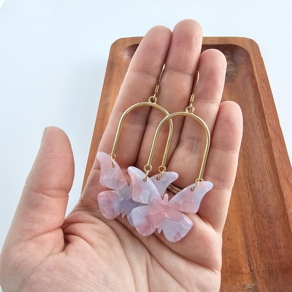 Hand holding butterfly earrings: gold hoops with pale pink layered butterfly charms