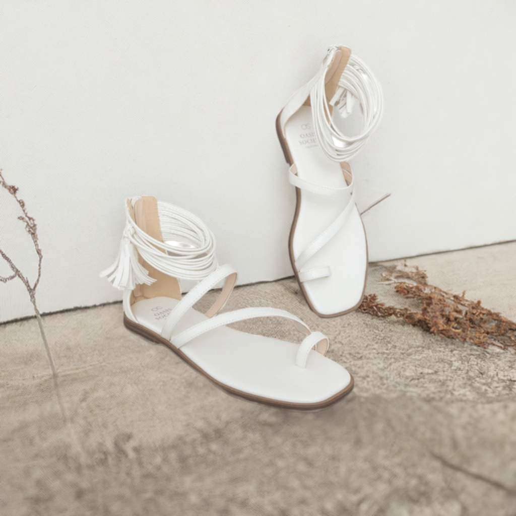 Pair of white ankle wrap flat sandals with multi-strap design and tassel fringe