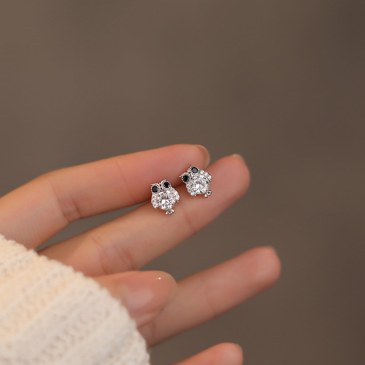 Pair of owl stud earrings shown in hand; silver with clear stones and black eyes