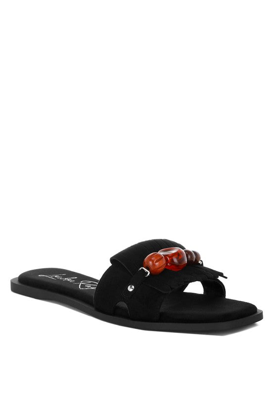 Black microfiber slip-on flat sandals with tortoise bead embellishment and fringe detail