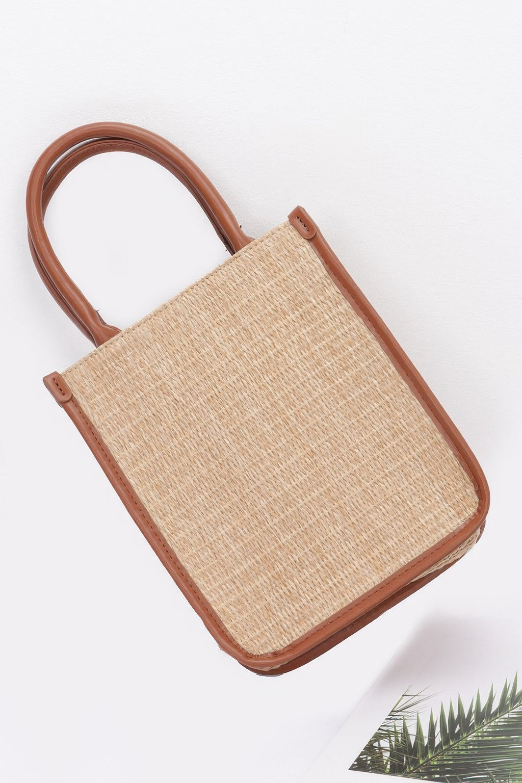 Diagonal view of khaki faux straw tote bag with tan leather trim and handle