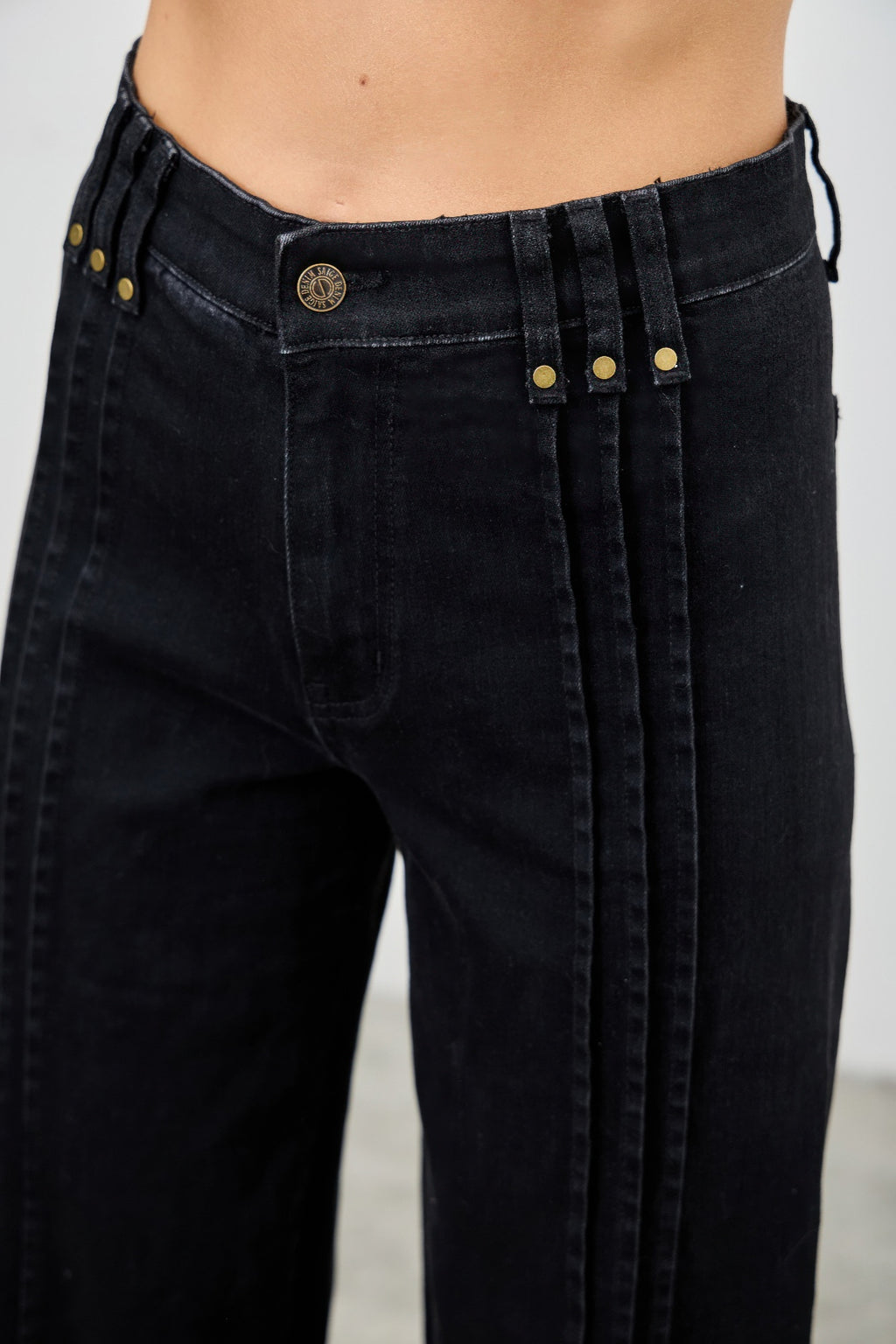 Close-up of high-rise black jeans front: round logo button and vertical seam stripes