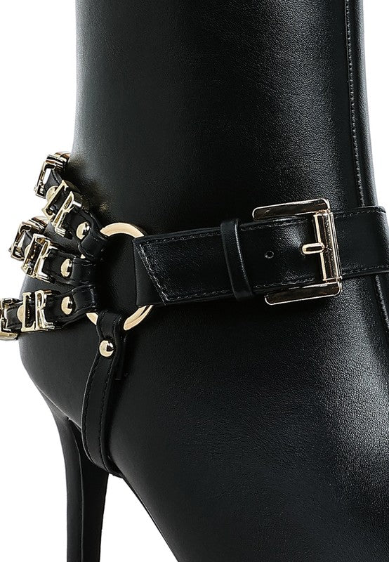 Black harness-style stiletto boot close-up with front belt strap and gold-tone studs