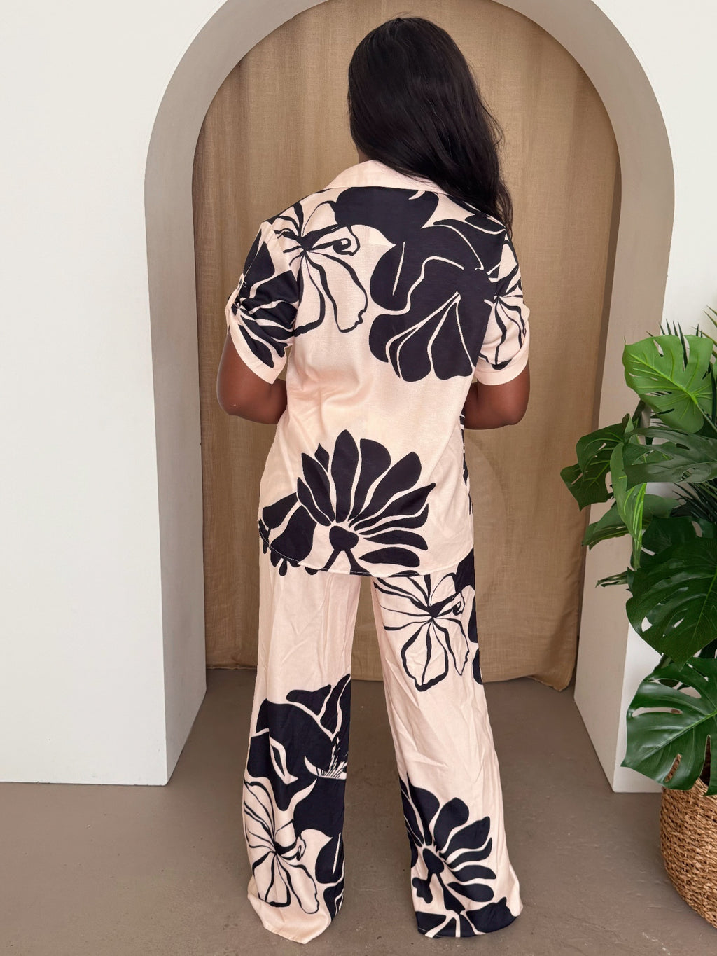Back view: cream pants set with black floral print, short-sleeve button-up top and wide legs