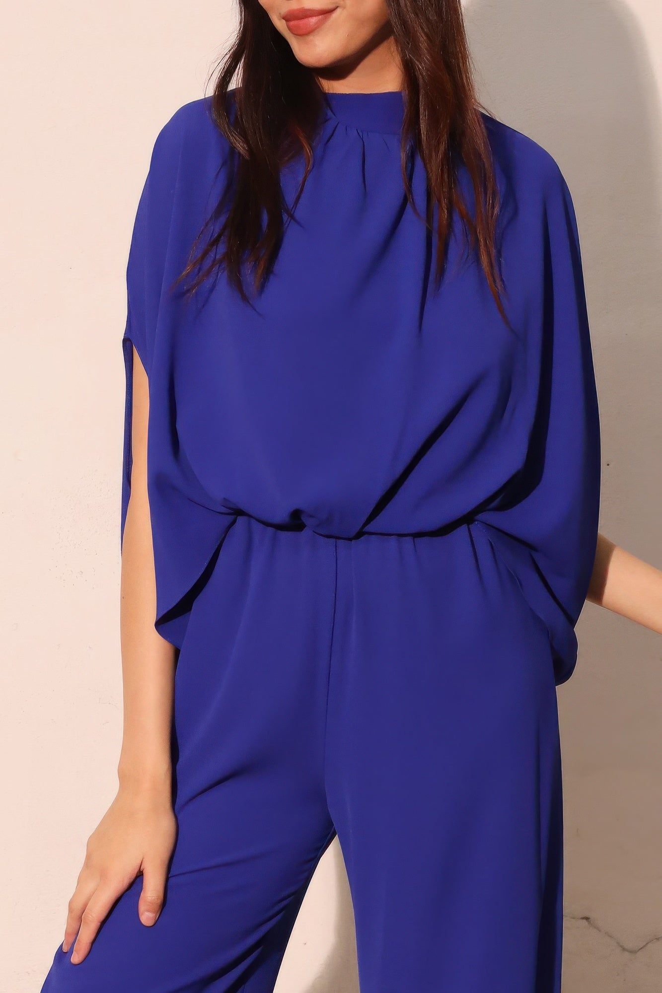Royal blue mock-neck jumpsuit with draped dolman sleeves and tie-waist detail.