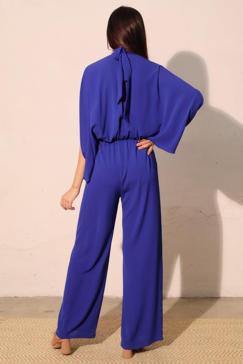 Back view: royal blue mock-neck jumpsuit with neckline tie and wide-leg pants.